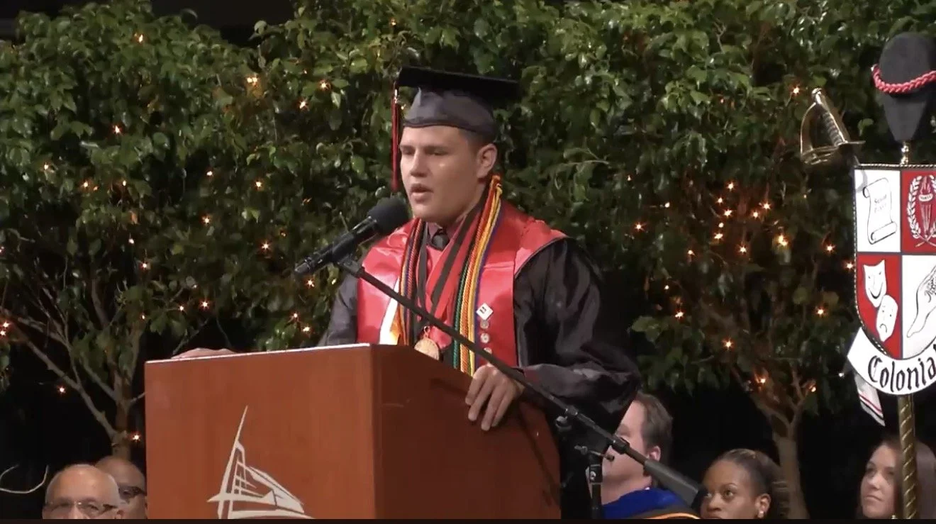A male graduate in a black cap and gown stands at a podium, speaking into a microphone during a graduation ceremony. Behind him are students and a banner with a crest and the word 'Colonia'. The background has greenery and string lights.