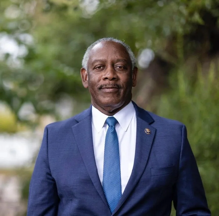 Portrait of a middle-aged African American man with gray hair, wearing a dark blue suit, white shirt, and blue tie, standing outdoors with blurred greenery in the background.