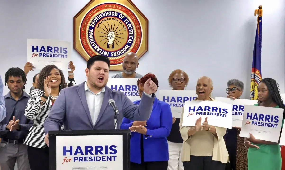 A group of diverse people at a political rally supporting Harris for President, with signs and a man speaking at a podium in front of a Brotherhood of Electrical Workers emblem.
