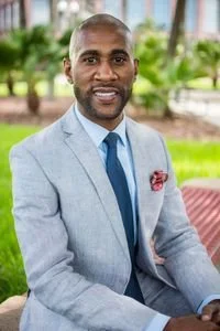 Portrait of a man wearing a light gray suit and blue tie, sitting outdoors with trees and grass in the background.