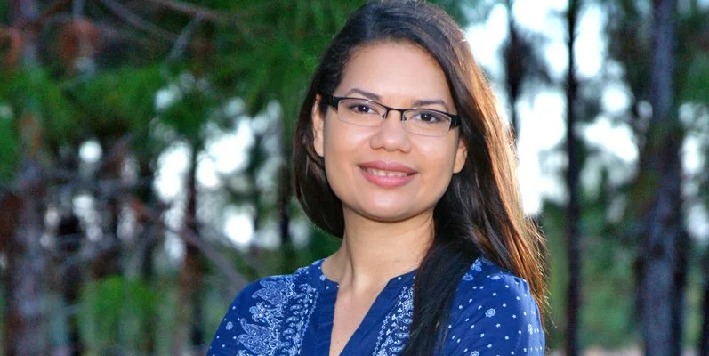 A woman with long dark hair and glasses smiling outdoors with trees in the background.