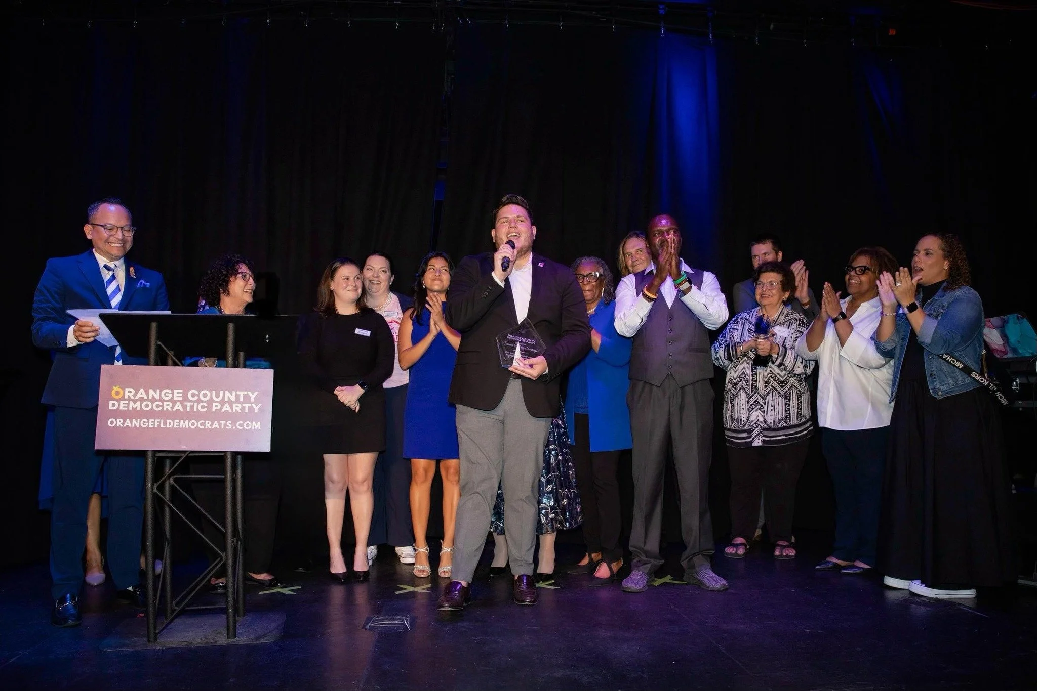 A group of people on stage at an Orange County Democratic Party event, with one person holding an award and speaking into a microphone, surrounded by supporters clapping and smiling.