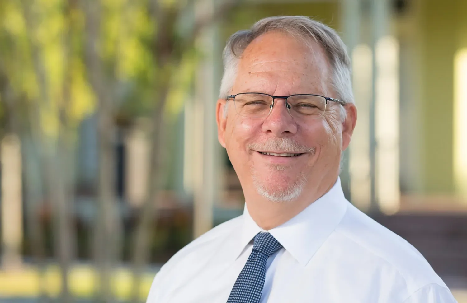 Photo of a smiling middle-aged man with glasses, wearing a white shirt and blue tie, outdoors with trees and a building in the background.