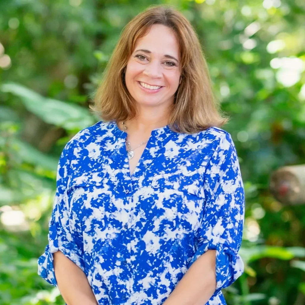 A smiling woman with shoulder-length light brown hair, wearing a blue and white patterned blouse, standing outdoors with greenery in the background.