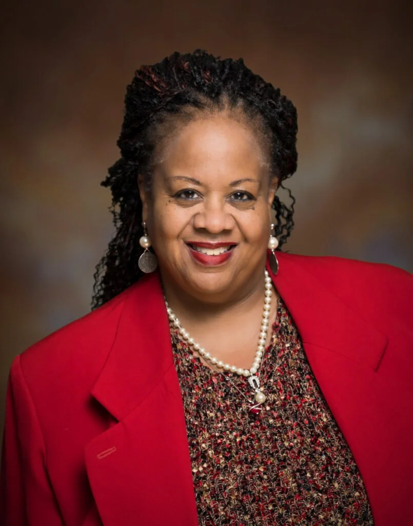 Portrait of a smiling Black woman with braided hair, wearing a red blazer, a patterned blouse, and pearl jewelry against a brown background.