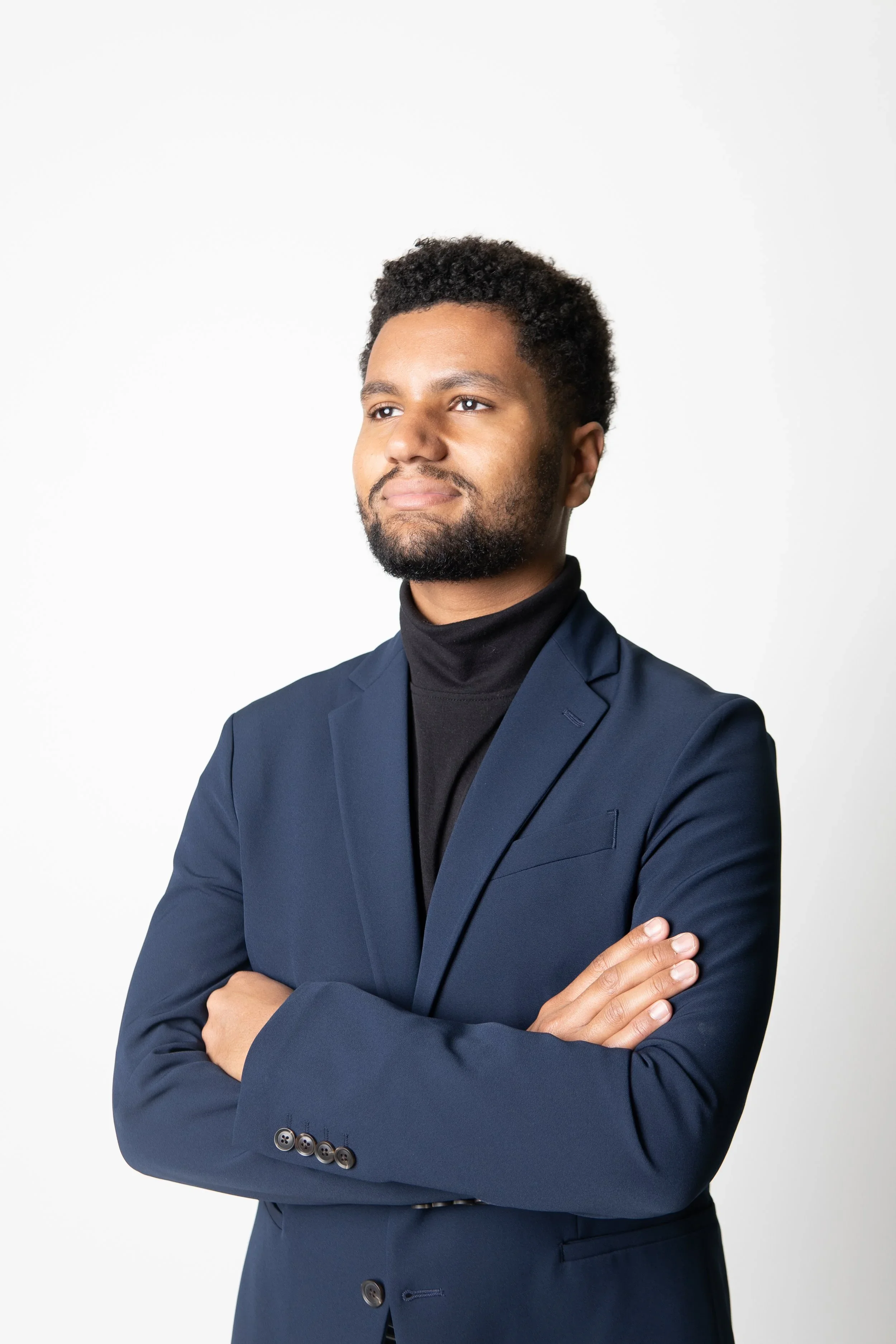 A young man with dark skin, short curly hair, and a beard, is wearing a black turtleneck and a navy blue blazer, standing with his arms crossed against a plain white background.