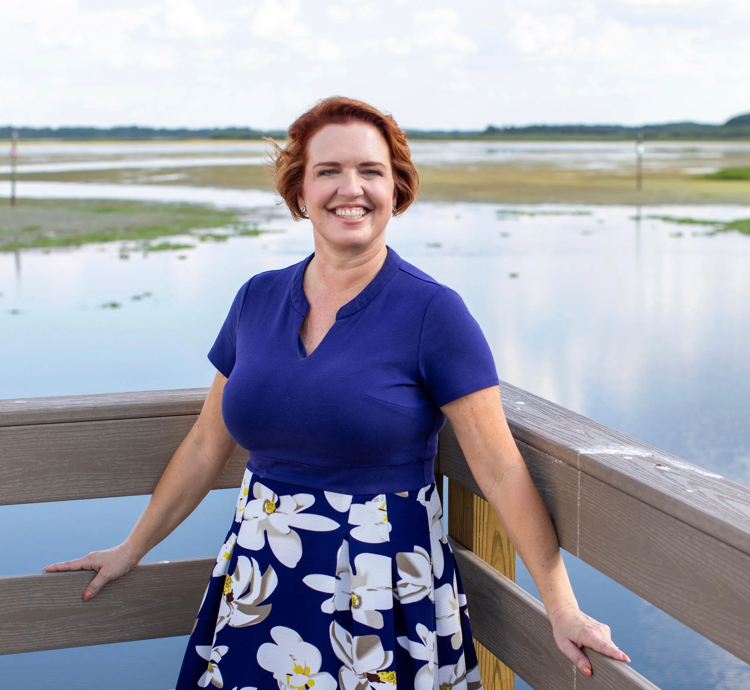 A woman with short red hair smiling and standing on a wooden railing outdoors, with a body of water and marshland in the background.