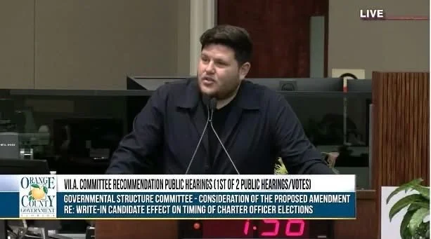 Man speaking at a podium during a public hearing in a government setting, with a digital clock and a plant nearby.