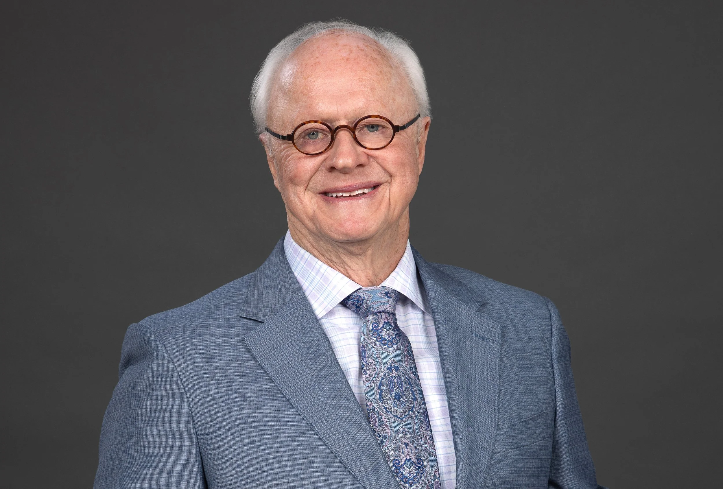 An elderly man with glasses and white hair smiling, wearing a light blue plaid suit, a patterned tie, and a collared shirt, against a dark gray background.