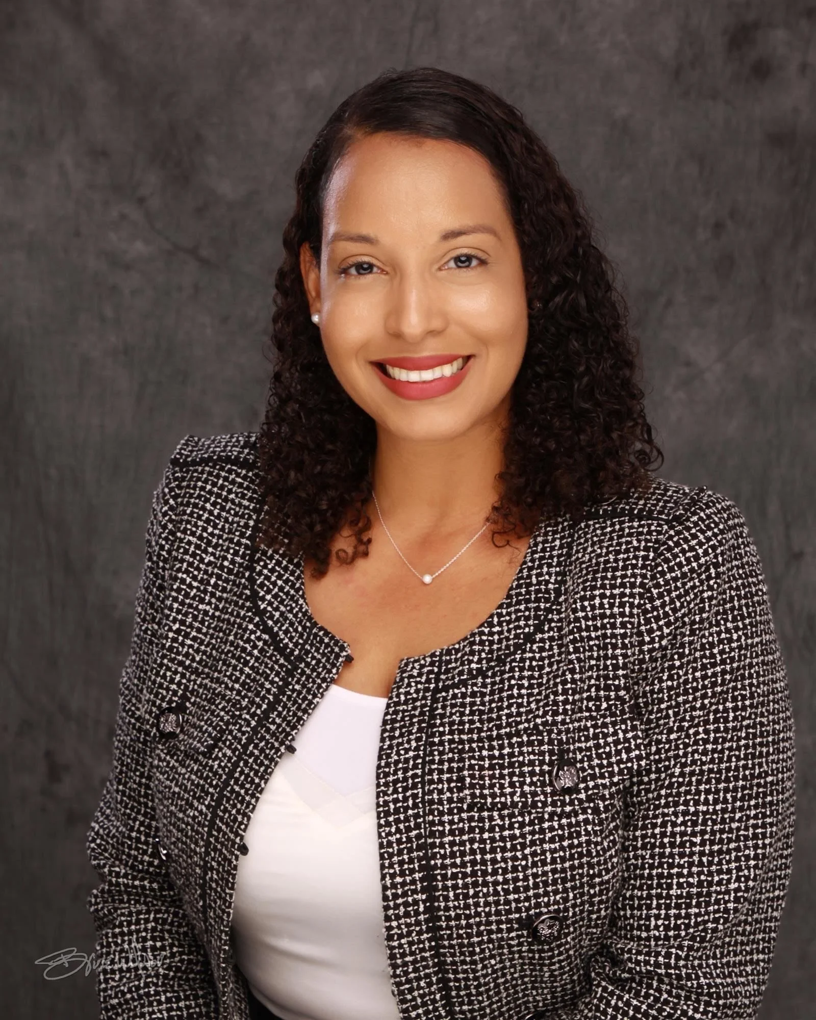 A professional woman with curly dark hair wearing a black and white checkered blazer and a white top, smiling at the camera against a dark, textured background.