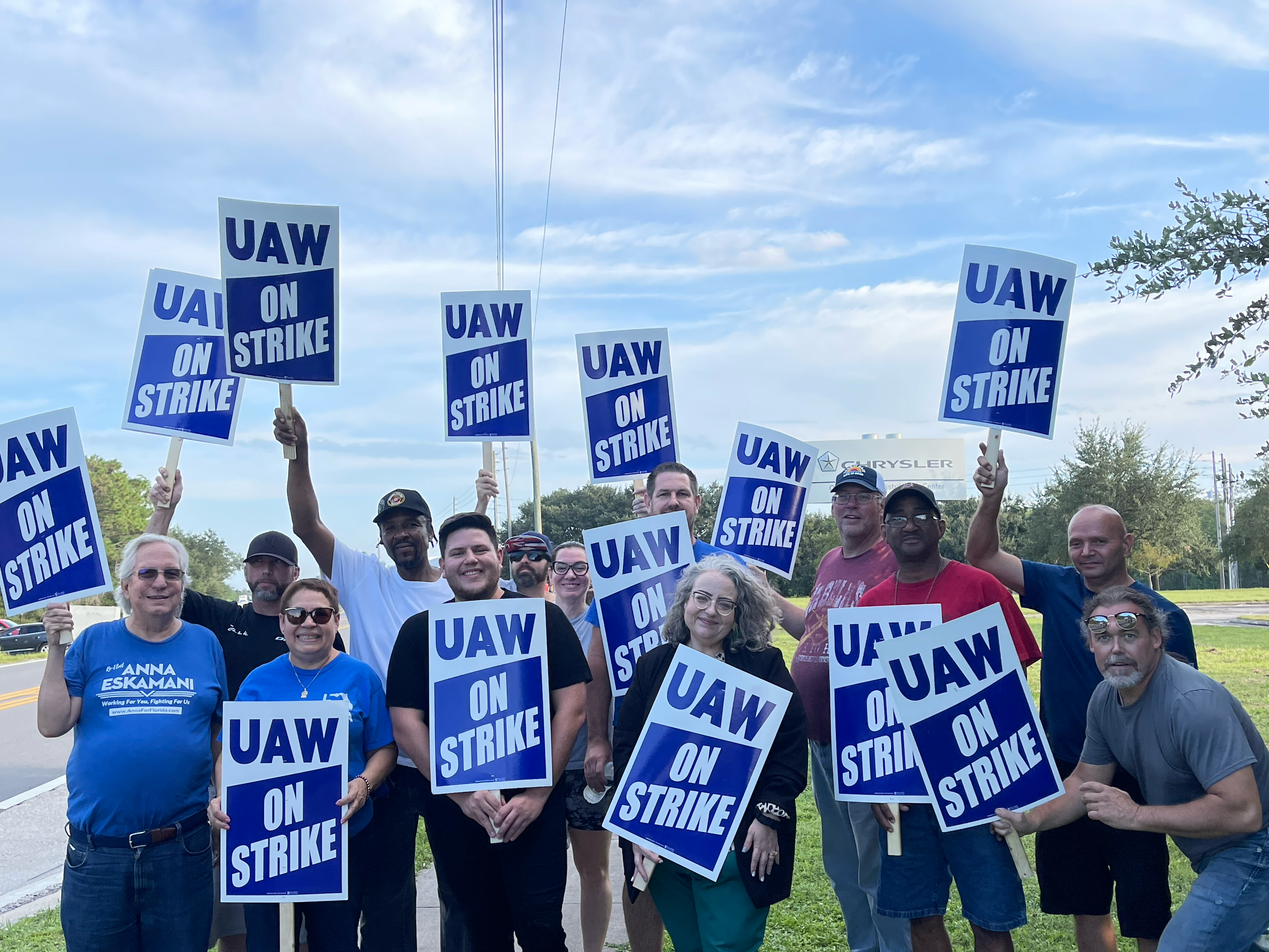 Group of people holding blue and white signs that read "UAW ON STRIKE" during a protest or strike event outdoors on a cloudy day.