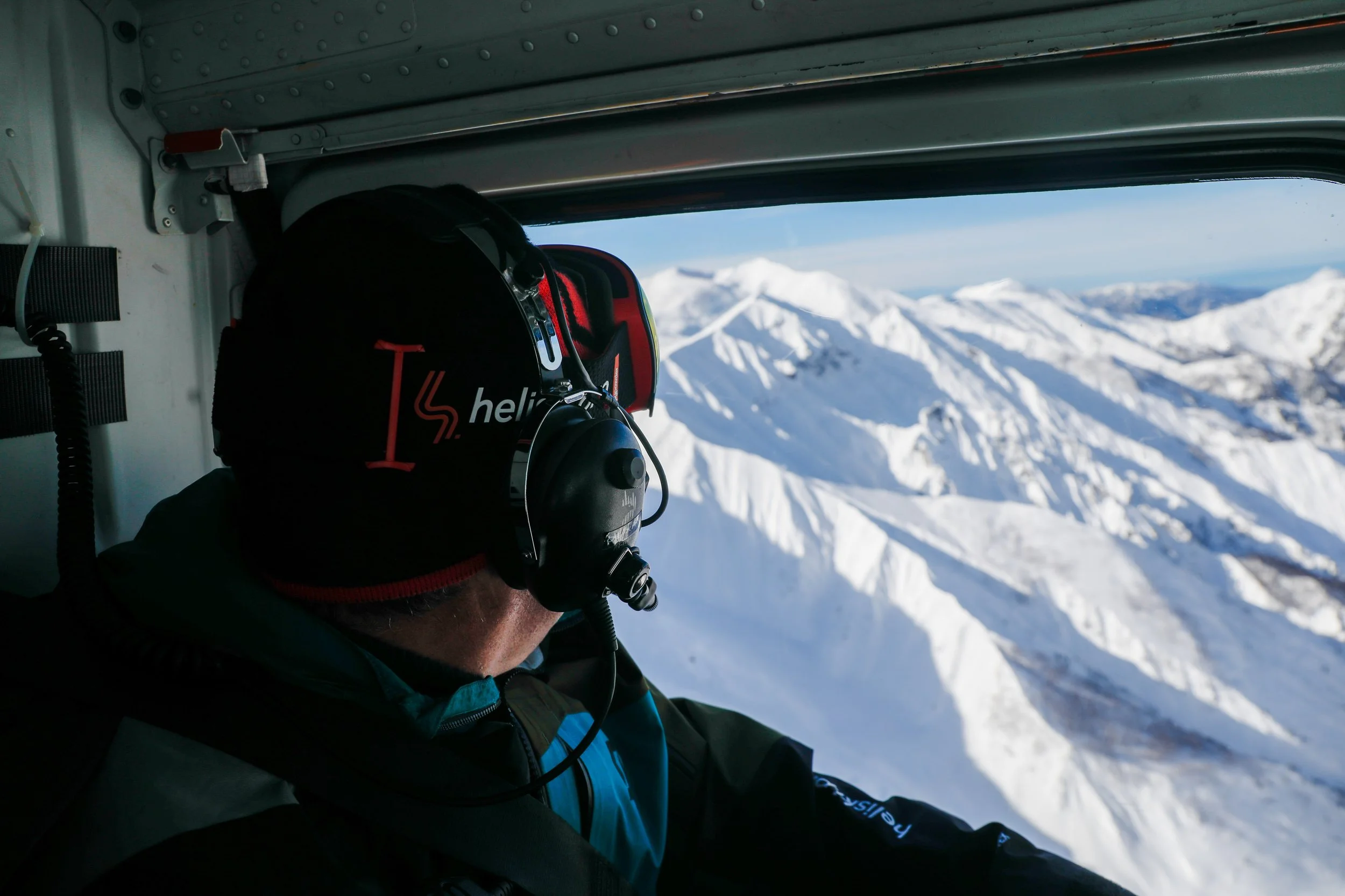 Una persona con cuffie e casco si affaccia da una finestra di un elicottero, con montagne innevate sullo sfondo.