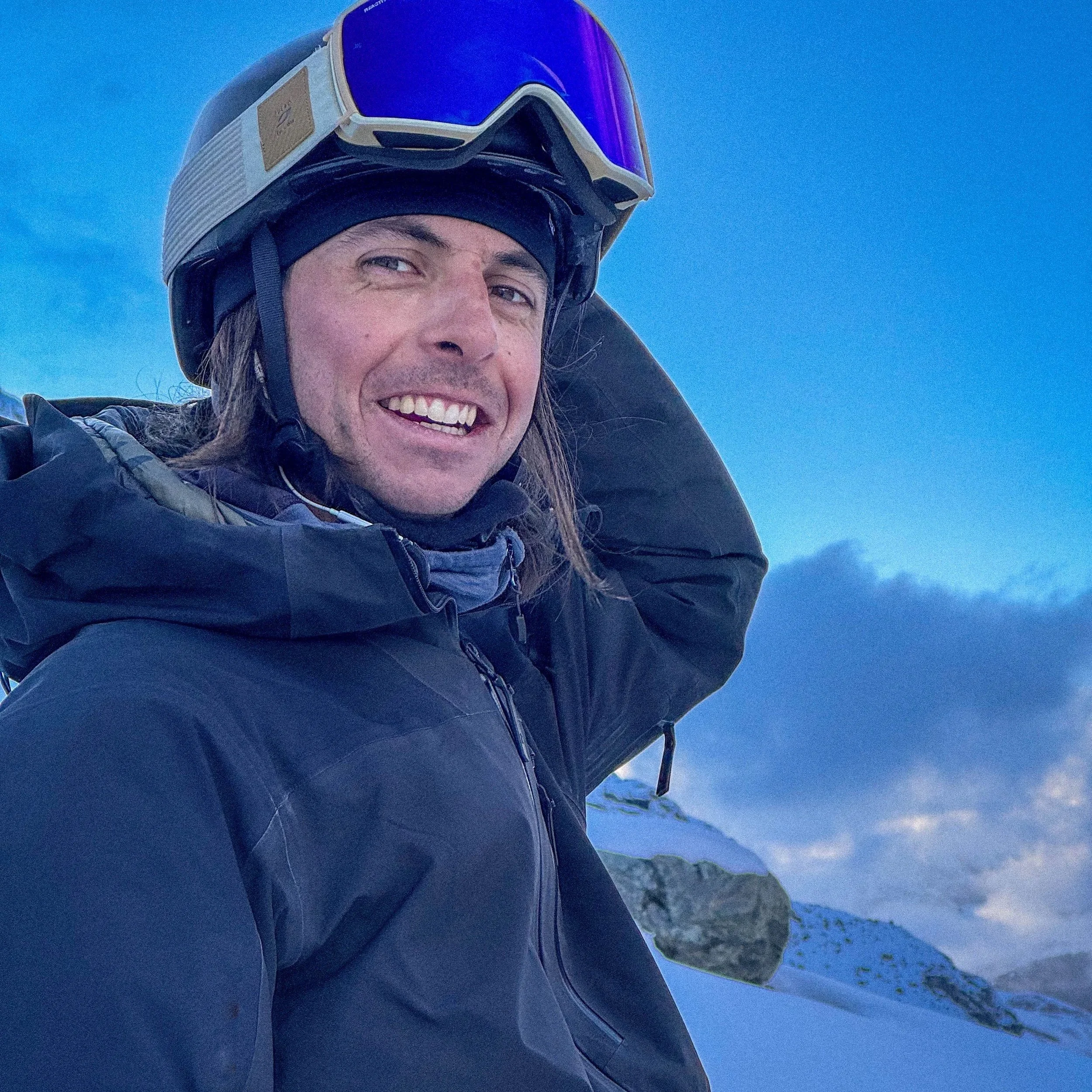 Uomo sorridente con casco da sci e giacca da montagna in un paesaggio ennevato e montano.