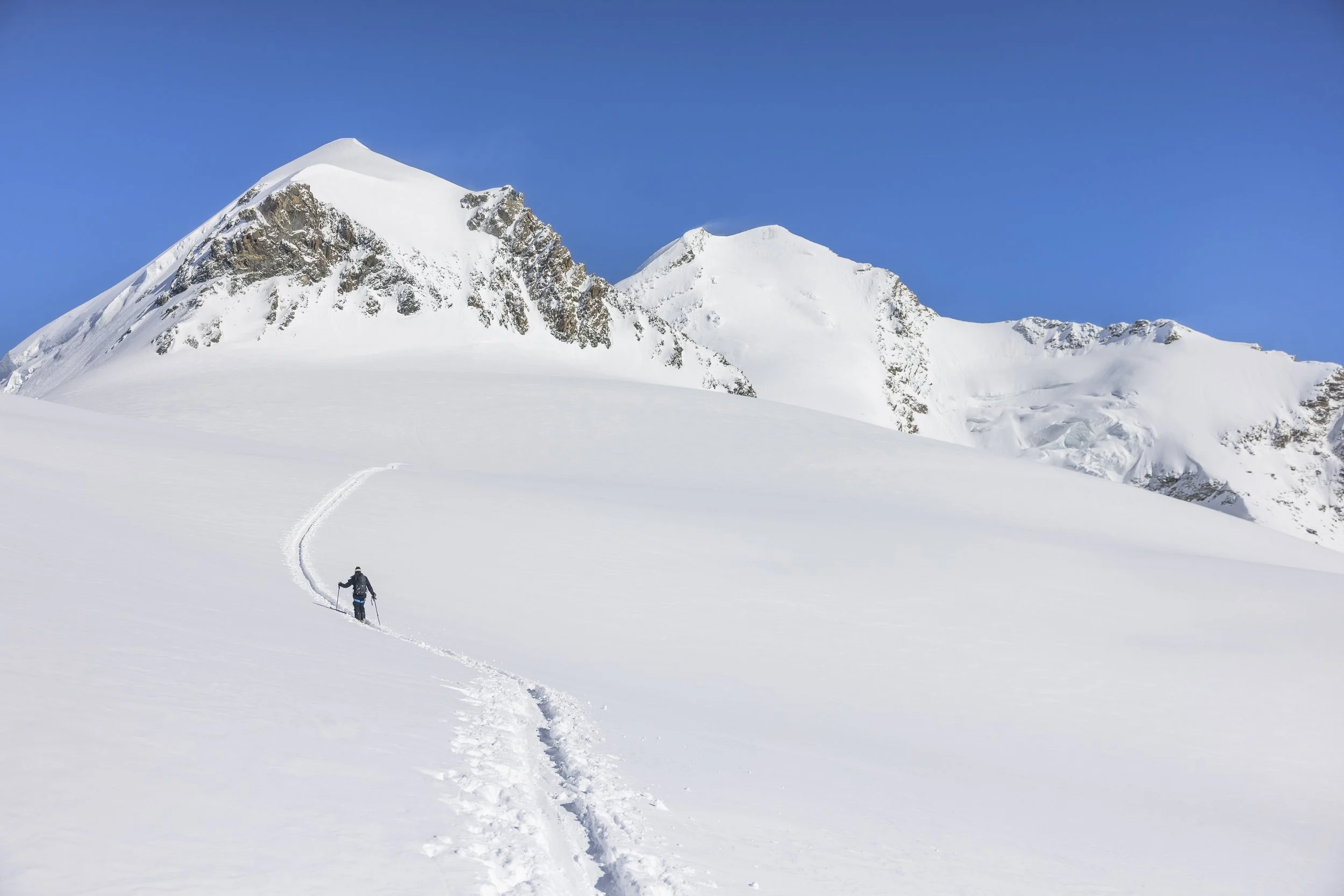 Una persona che fa trekking sulla neve, con montagne innevate sullo sfondo e un cielo azzurro.