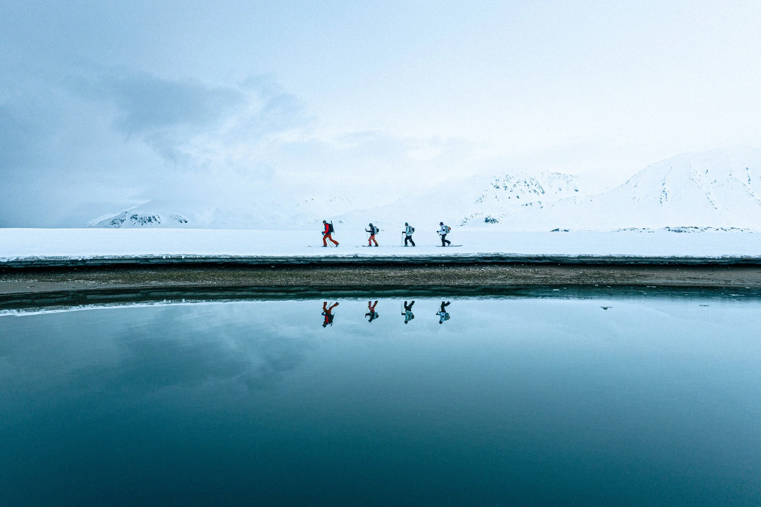 Cinque persone che camminano nella neve in un paesaggio glaciale, con montagne in lontananza, e il loro riflesso in un lago calmo.