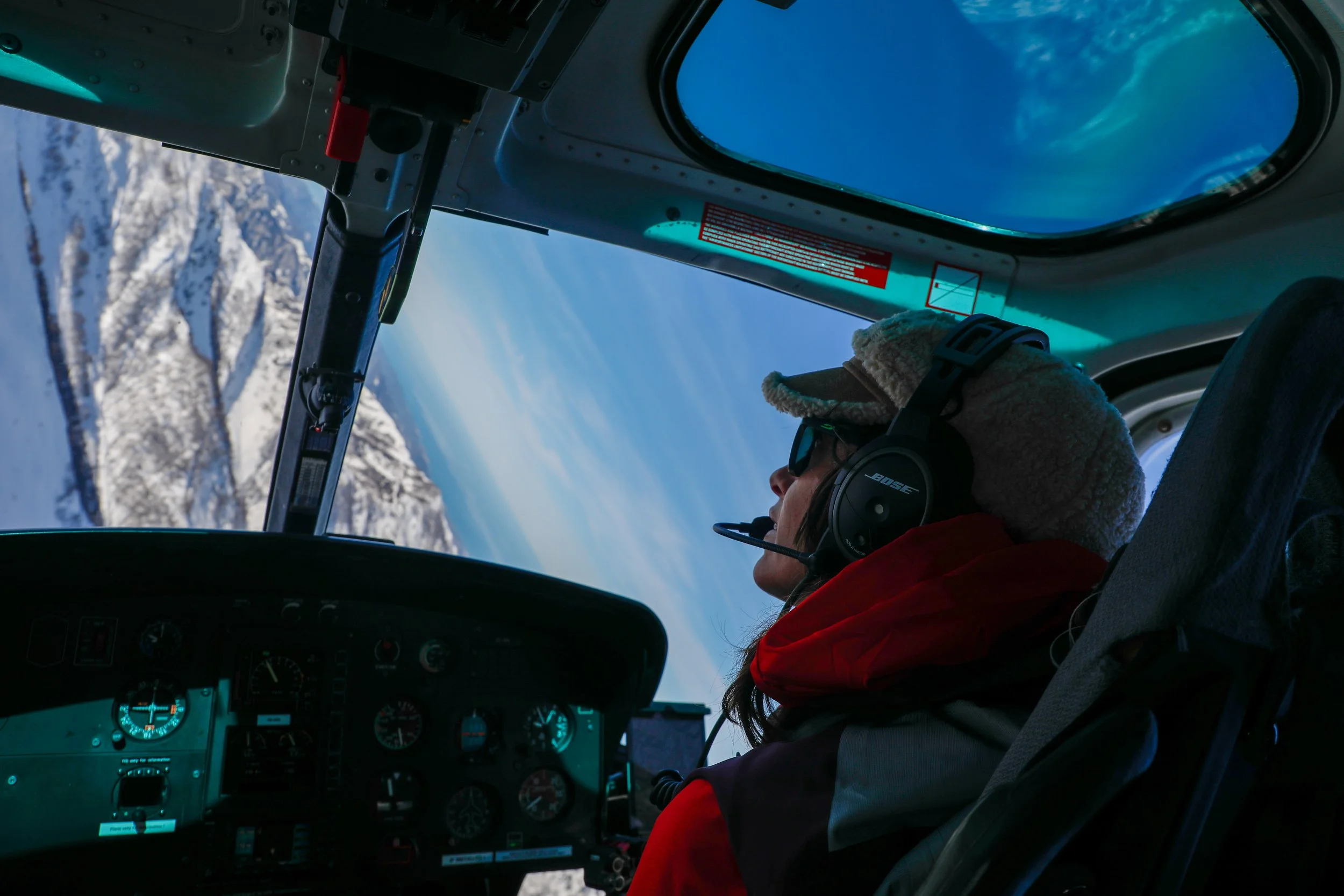 Pilota in cabina di un elicottero che sorvola le montagne innevate con cielo blu.