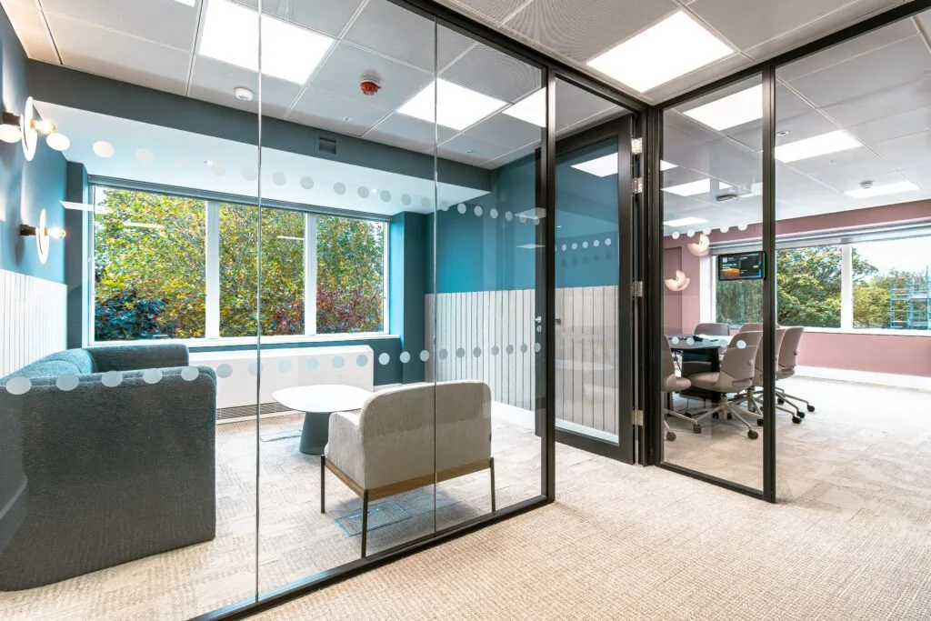 Office meeting room with glass walls, seating area with sofa and chairs, and a conference table with chairs; large windows showing trees outside.