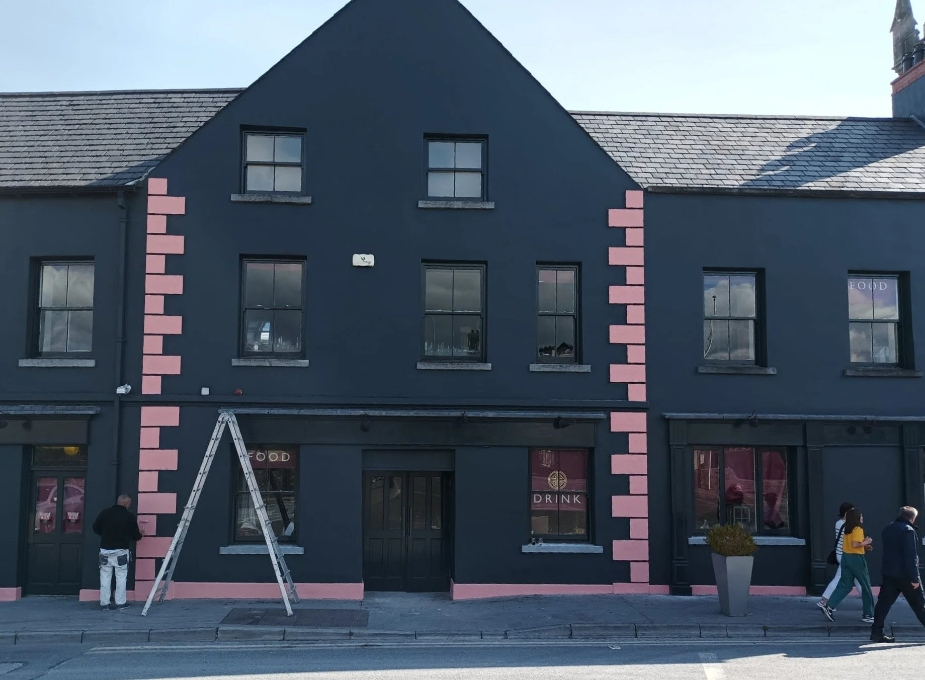 Building with dark exterior, pink decorative accents, and multiple windows; people walking on sidewalk in front.