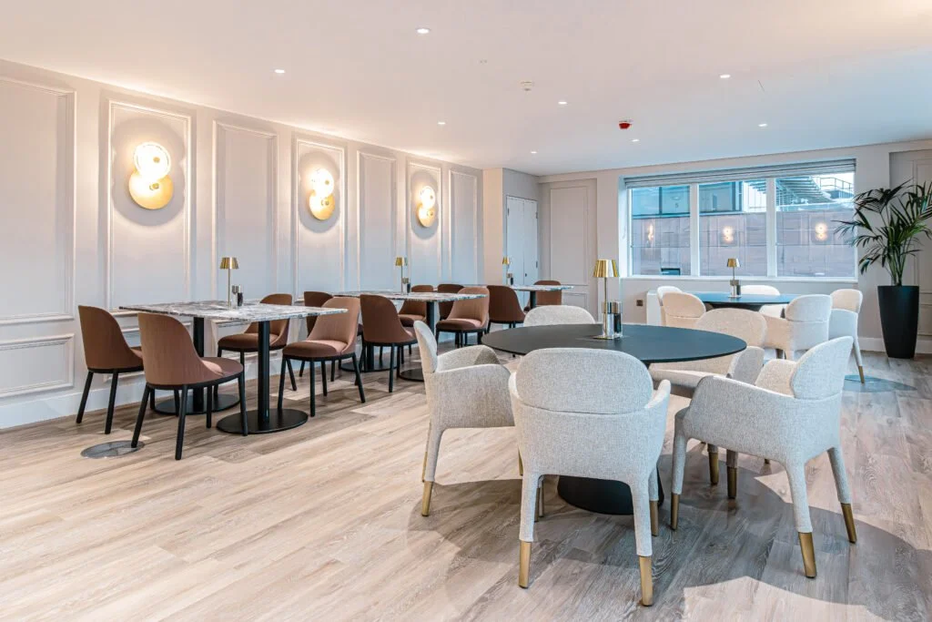 Modern dining area with round tables, beige and pink chairs, wall sconces, and a large window with a view of neighboring buildings.