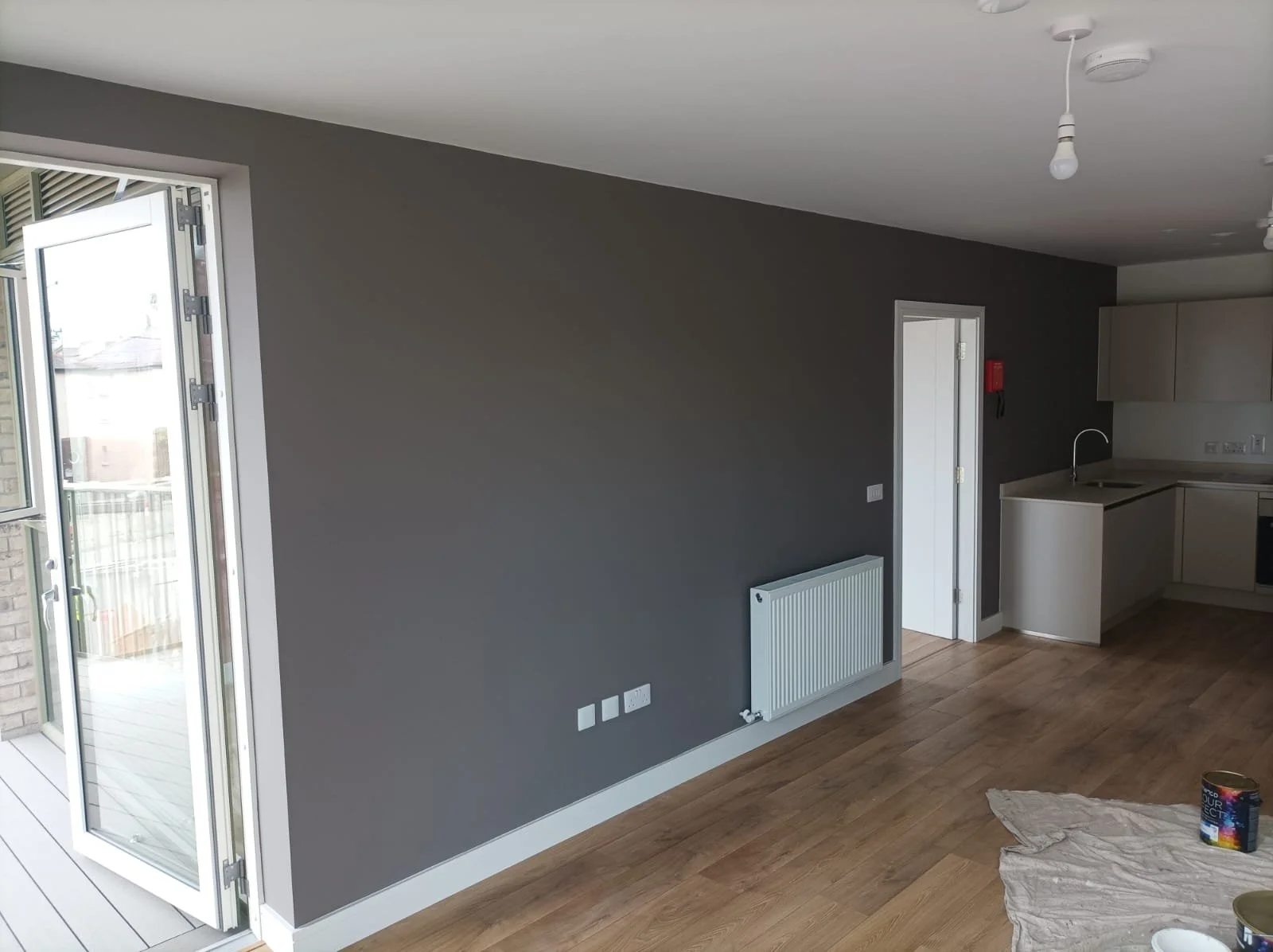 Interior of an unfurnished room with gray walls, a radiator, and a kitchen area with beige cabinets. An open door leads to a balcony or outdoor area, and a plastic sheet with a paint can is on the wooden floor.