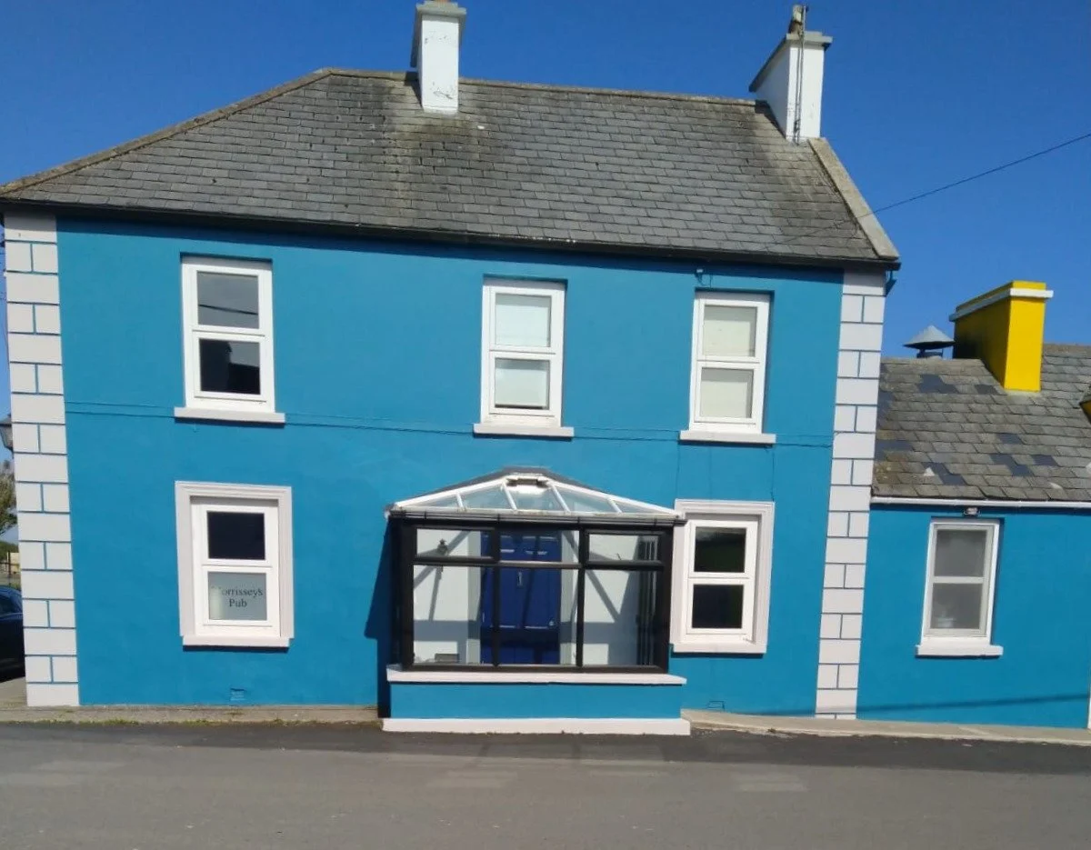 A blue, three-story building with white window frames and a glass-enclosed porch in the center. The building is on a street with a clear blue sky overhead.