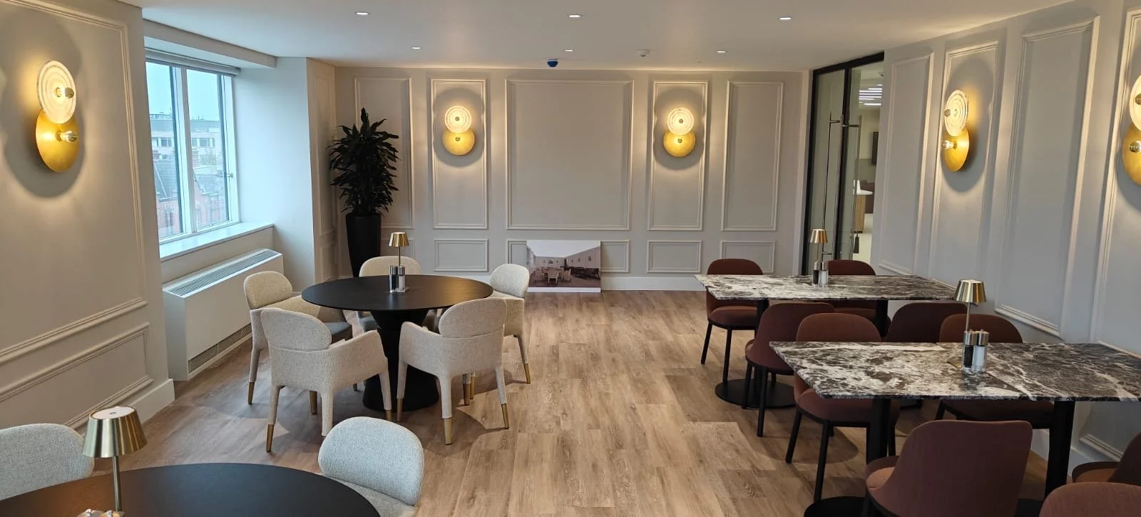 A modern dining area with round tables, beige and burgundy chairs, wall sconces, and a large window with city view.