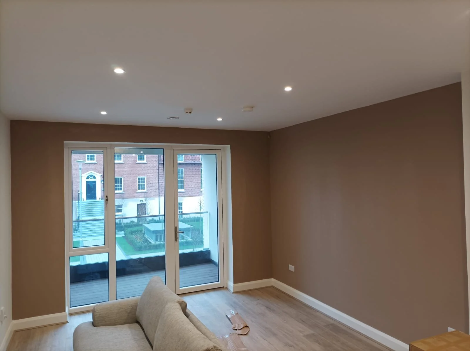 Empty living room with beige walls, wooden flooring, and glass door leading to a balcony, with a partial view of a sofa.