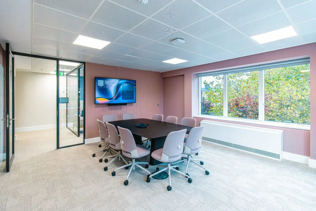 A modern conference room with a large black table surrounded by nine pink cushioned chairs on casters. There is a flat screen monitor mounted on a pink wall, and a large window with a view of trees outside. The room has a ceiling with white tiles and fluorescent lighting, beige carpet, and glass doors leading to another area.