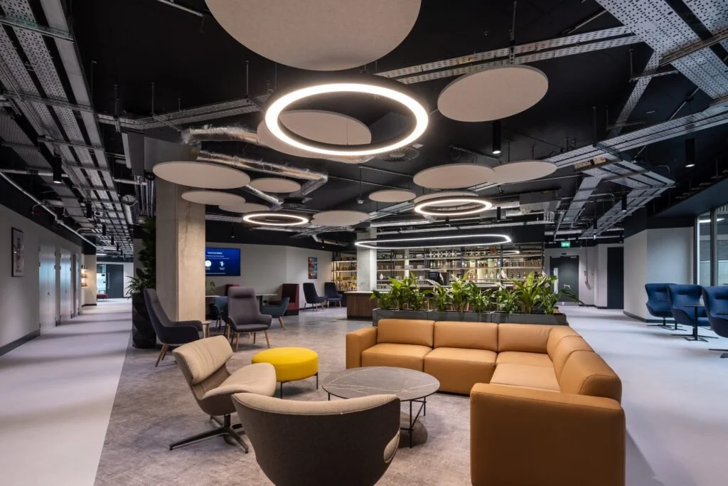 Modern office lounge area with beige sectional sofa, grey and yellow chairs, circular coffee table, and indoor plants. Overhead circular and linear LED light fixtures, open ceiling with exposed ducts, and a bar with shelves in the background.