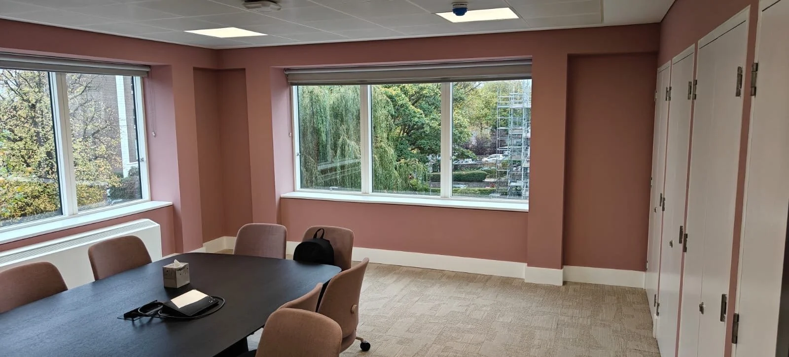 Room with pink walls, large windows showing outdoor trees and scaffolding, a black table with pink chairs, a tissue box, a notebook, and a bag, white cabinets on the right side.
