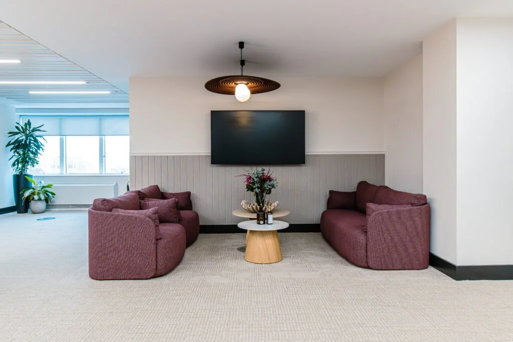 Modern lounge area with two burgundy sofas facing each other, a small round table with a floral arrangement in the middle, a flat-screen TV on the wall, and a ceiling light fixture.