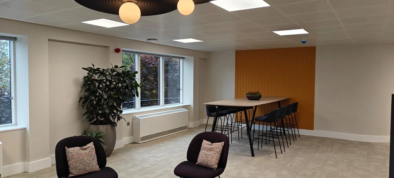 Modern office or lounge area with large window, potted plant, black chairs with patterned pillows, and a high table with chairs against a wood-paneled wall.