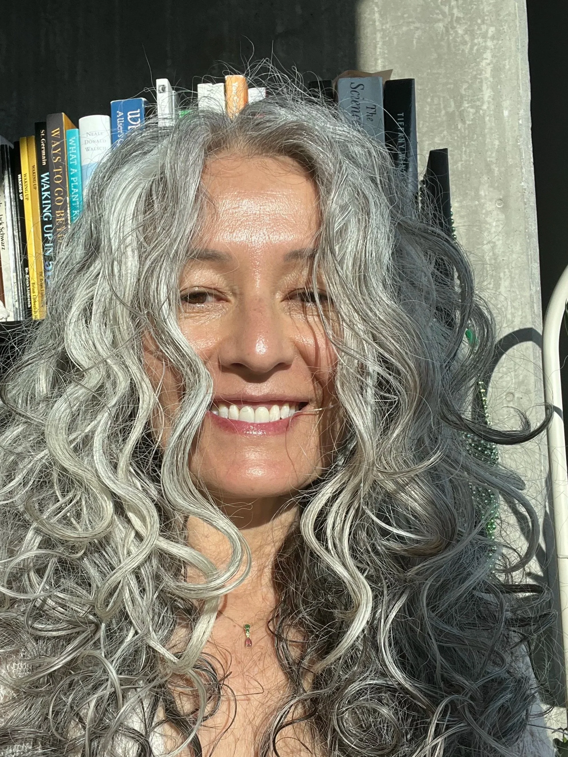 Close-up of a smiling woman with long curly gray hair, wearing a necklace with a small pendant, in front of a bookshelf with books. Sound Healer.
