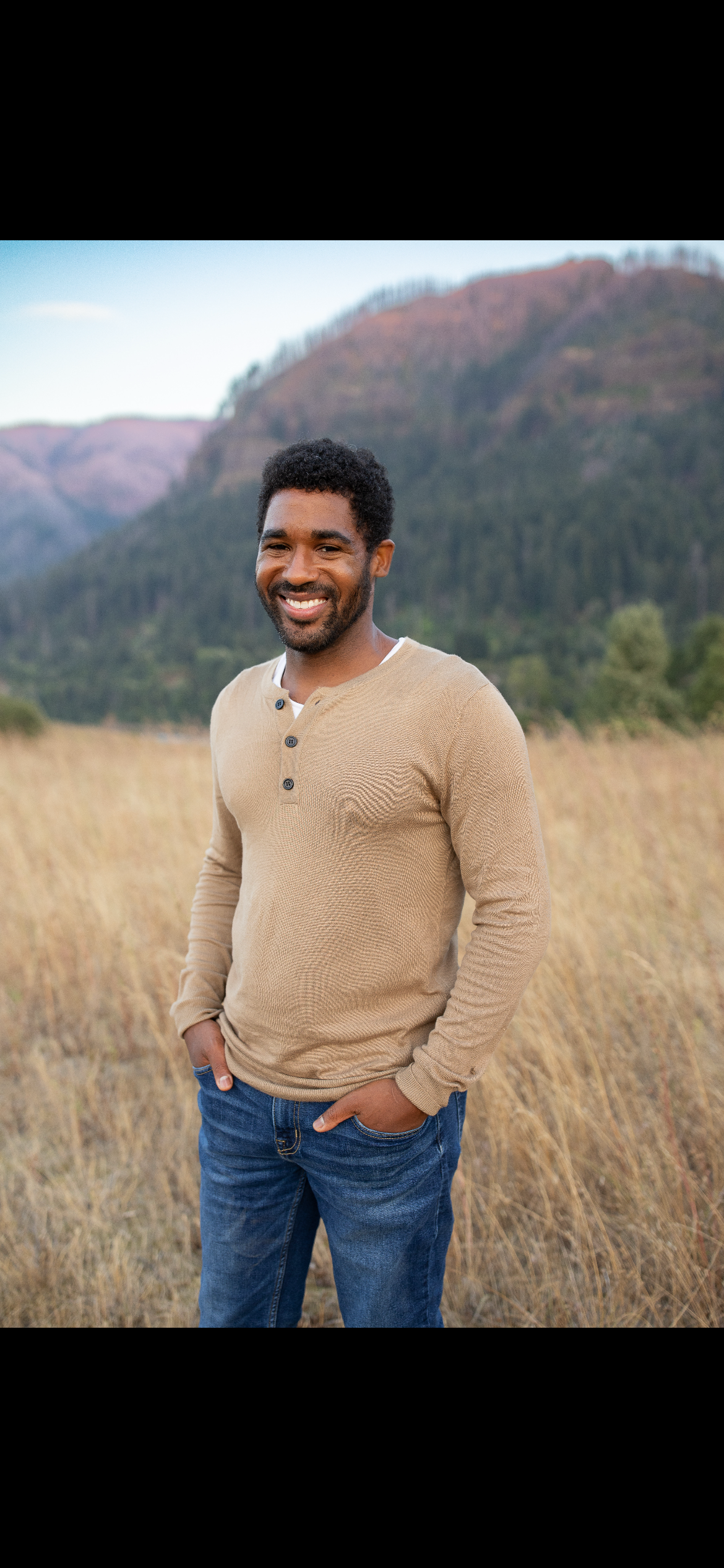 Smiling man standing outdoors in a field with mountains in the background. Sound Healer.