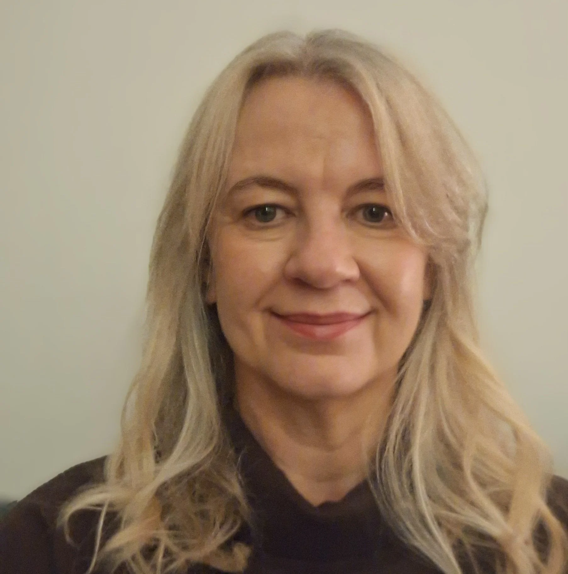 A woman with long blonde hair and a black shirt, smiling, in front of a plain light-colored wall.