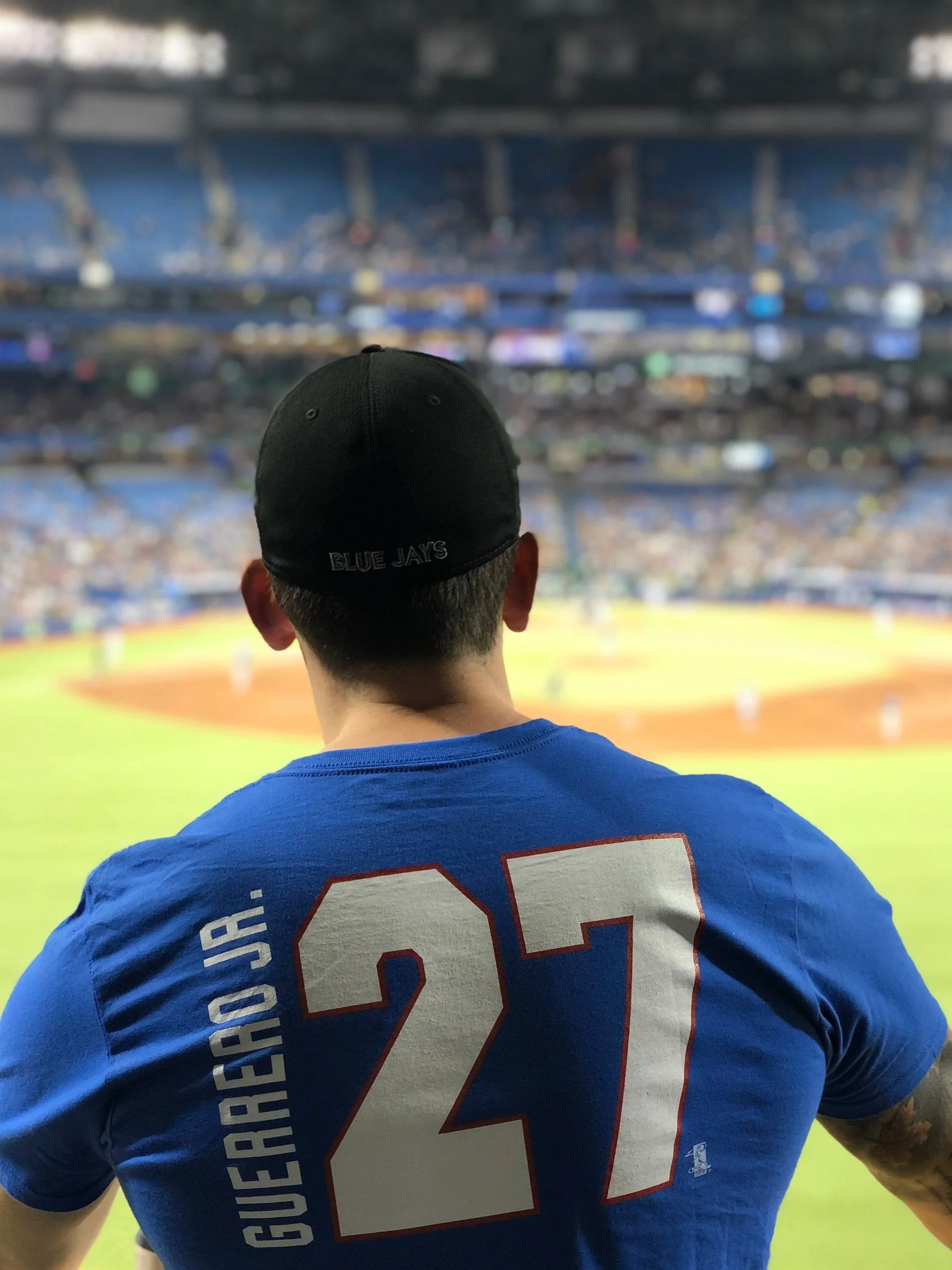 A baseball player wearing a blue jersey with the name 'GURRERO JR.' and the number '27' on the back, sitting in the stands and watching a game at a stadium.