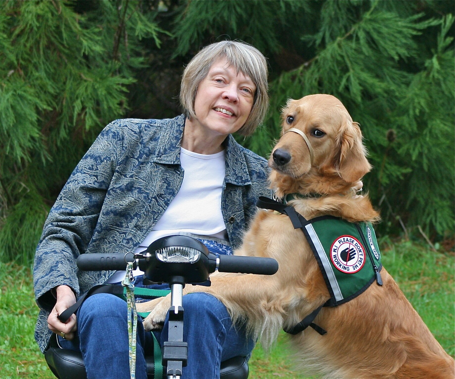 Karen, white women, sits in a motorized scooter outside. Golden retriever Narnia stands to Karens left, back paws on ground, front paws on Karens left leg.
