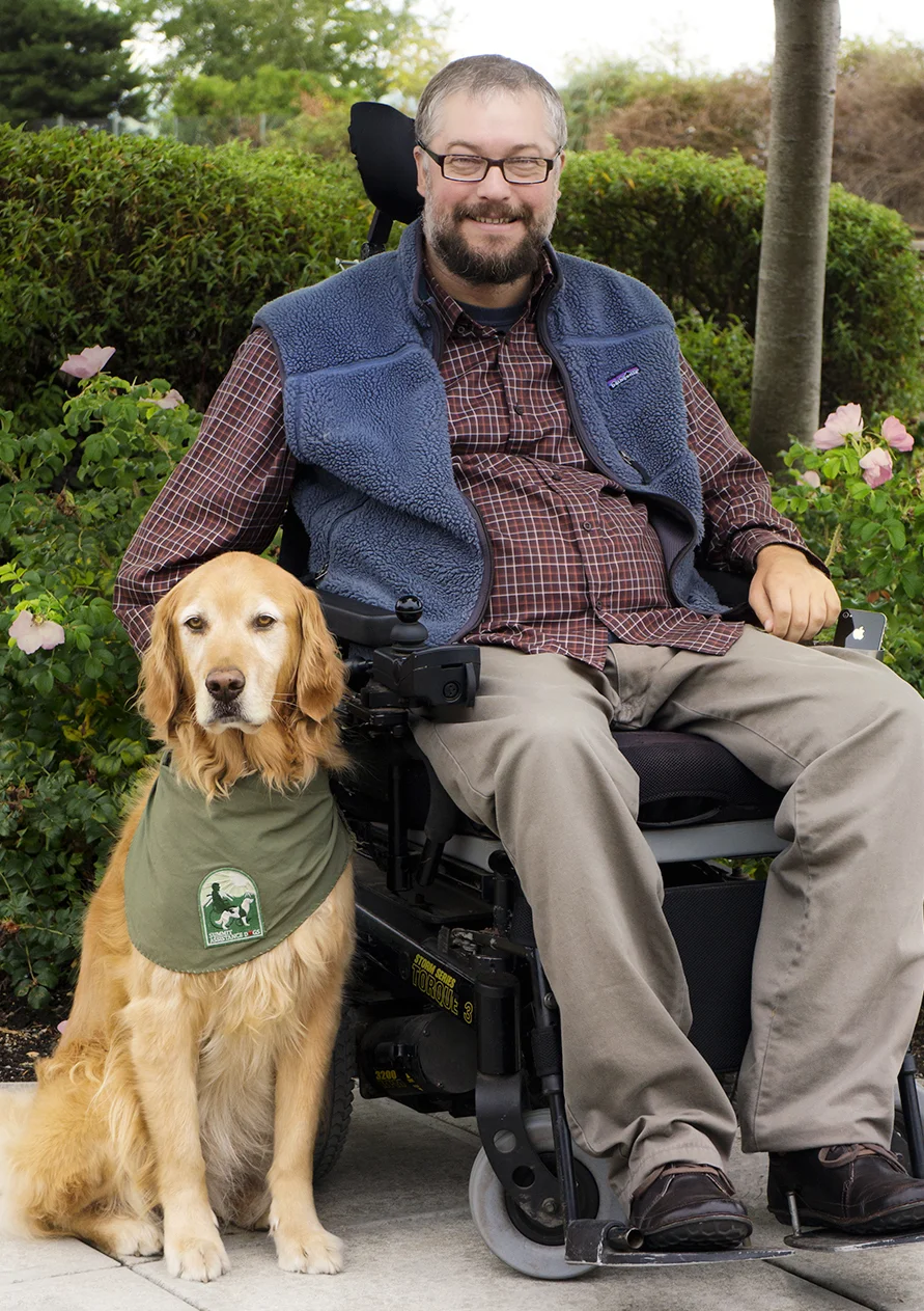 Eric sits in a motorized wheelchair facing camera and smiling. Titus, golden retriever, sits to the right of Eric, facing camera.