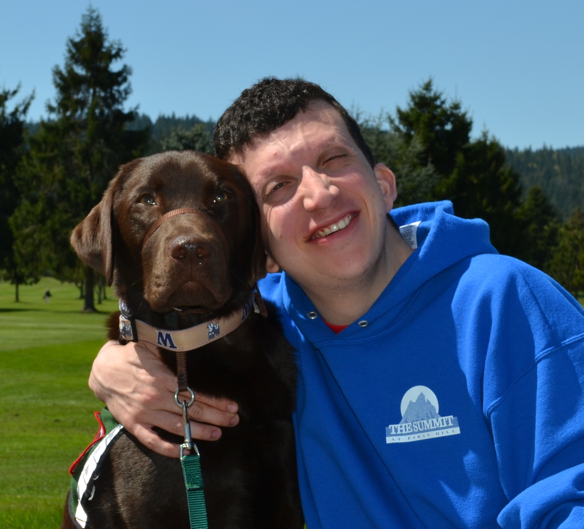 A young white man Alex, and chocolate labrador Baylor sit on grass together. They are facing the camera and resting their heads on each other.