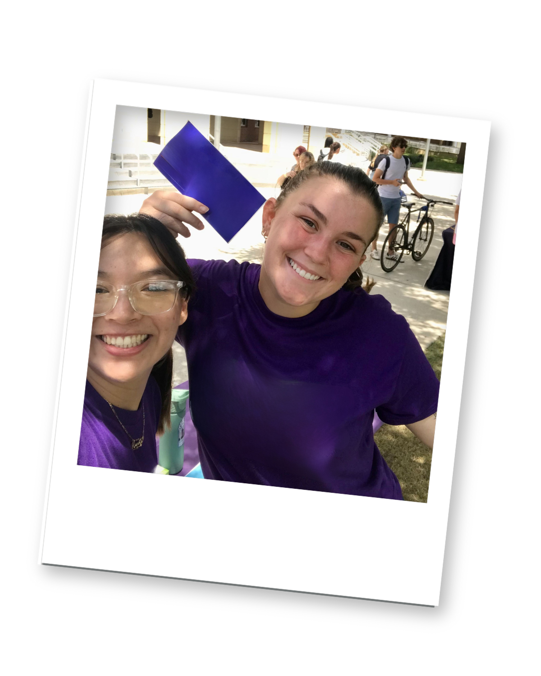 Two young women smiling, one with glasses and the other holding a blue diploma, outdoors on a sunny day.