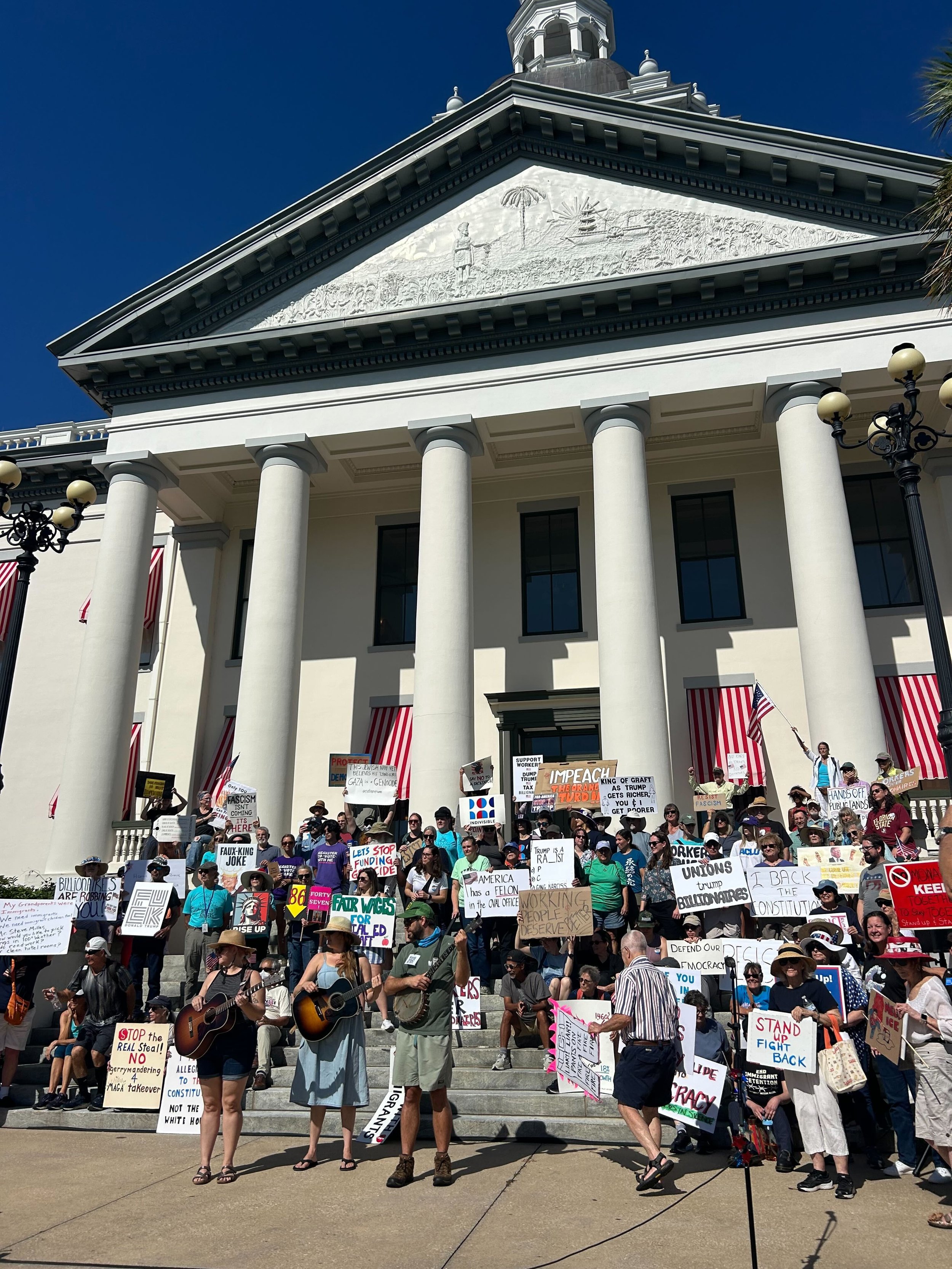 A group of protesters gathered on the steps of a government building, holding signs and banners with various messages, some playing guitars, under a clear blue sky.
