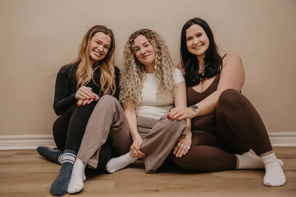 Three women sitting on the floor against a beige wall, smiling at the camera.
