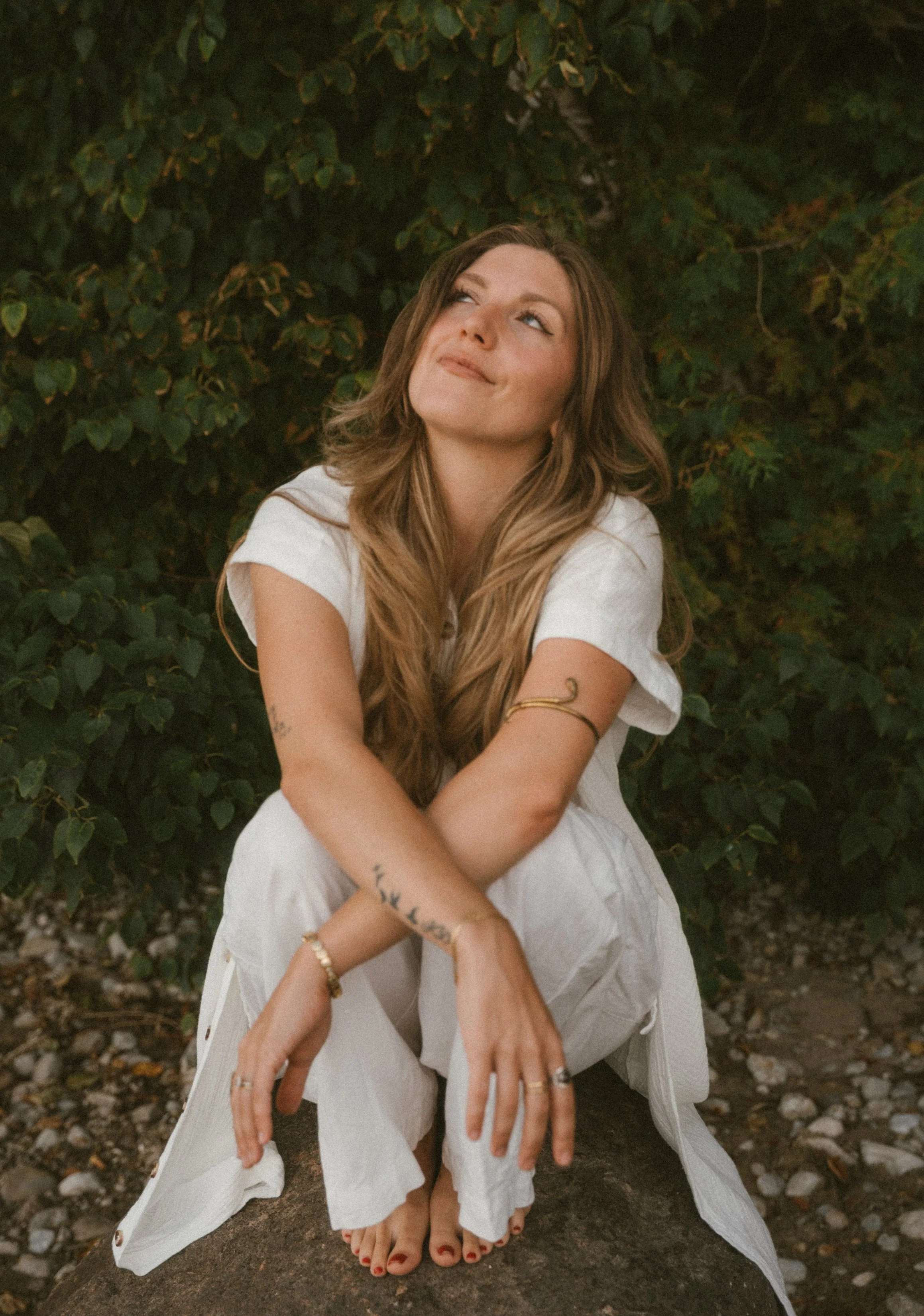 A woman with long wavy hair sitting on a rock outdoors, surrounded by greenery, looking up thoughtfully.