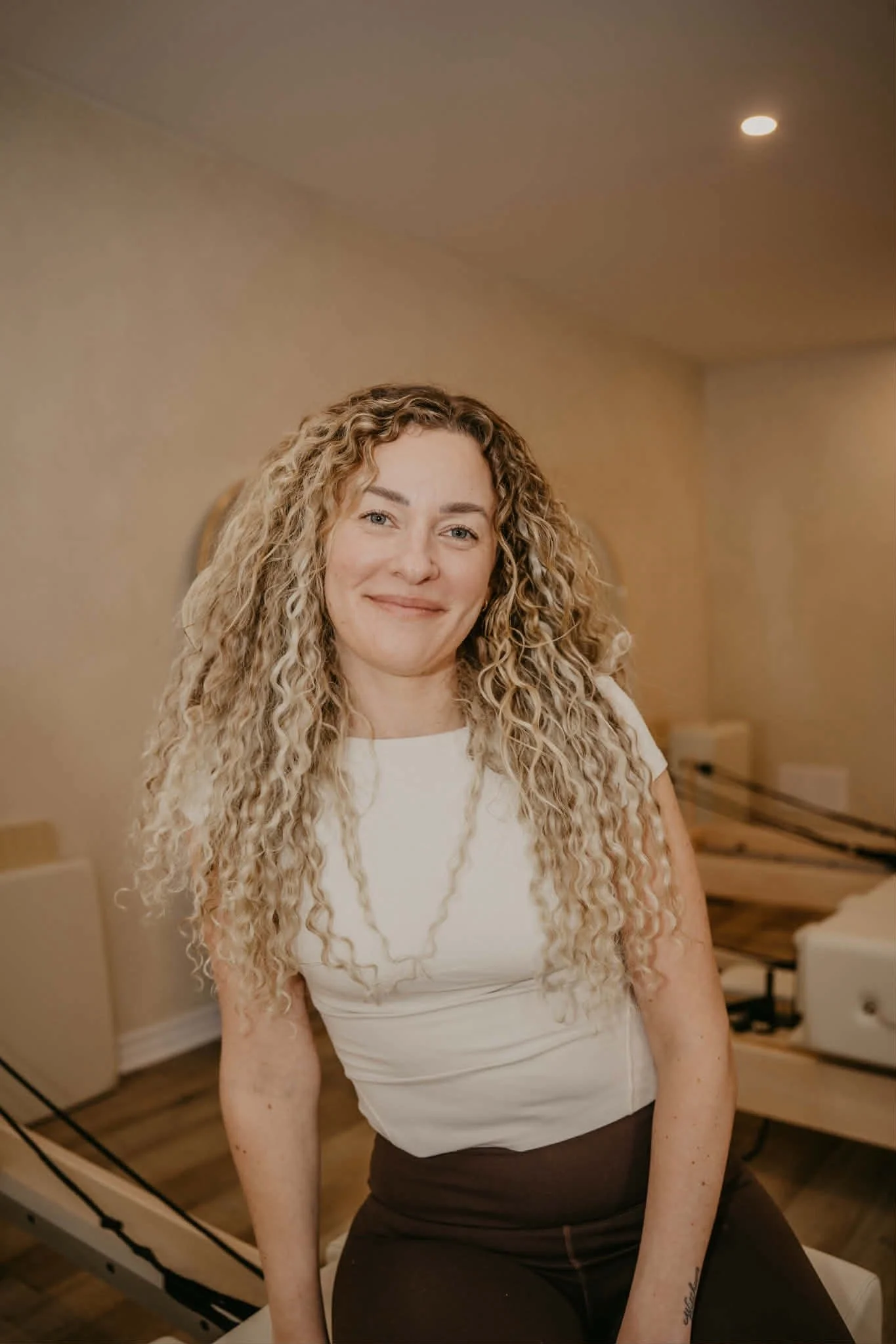 A woman with long, curly blonde hair smiling and sitting indoors, wearing a white sleeveless top and dark pants, in a warmly lit room.