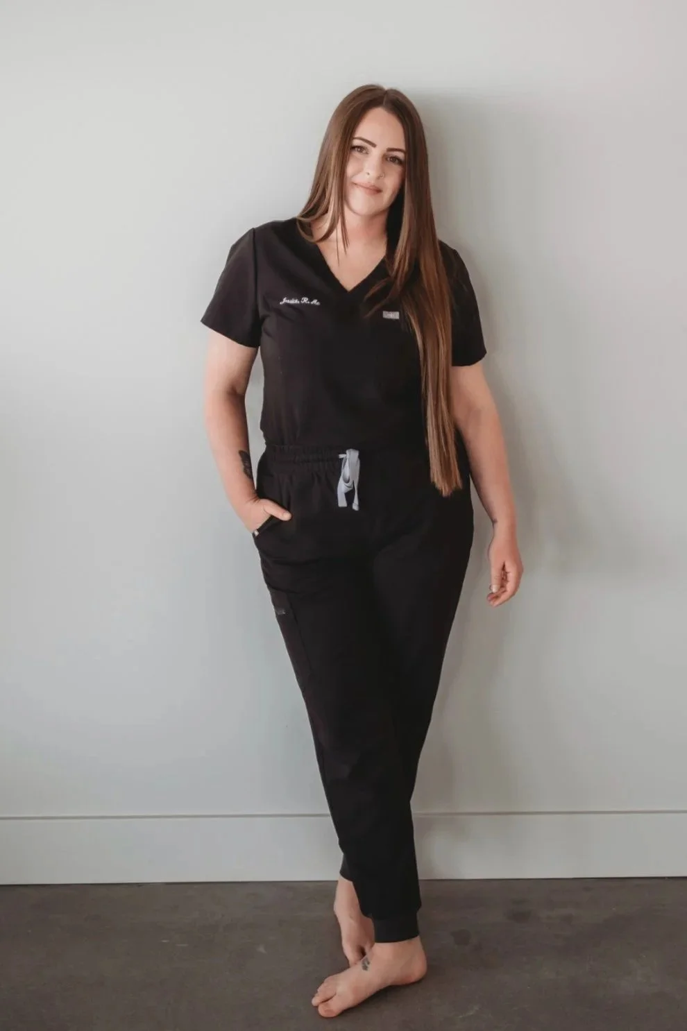 A woman with long brown hair in black medical scrubs, standing barefoot against a plain wall. She has her left hand in her pocket and is smiling slightly.