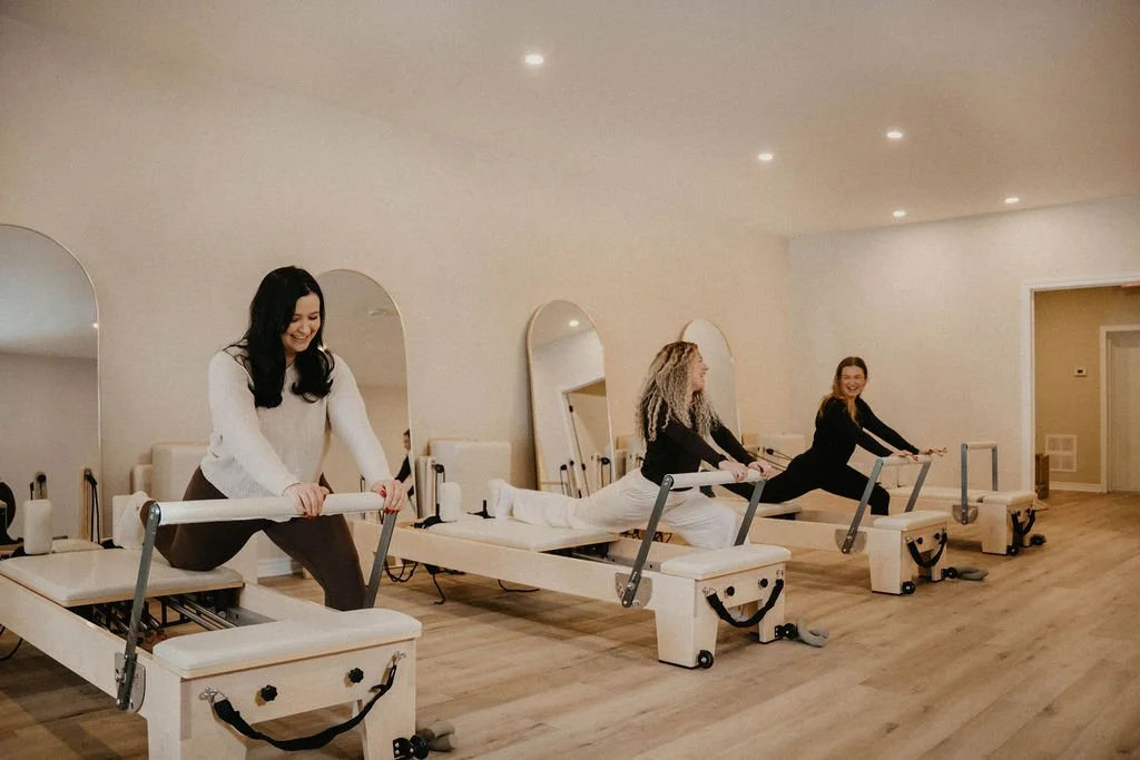 Three women on Pilates reformer machines in a bright, minimalistic studio, smiling and engaging in exercise.