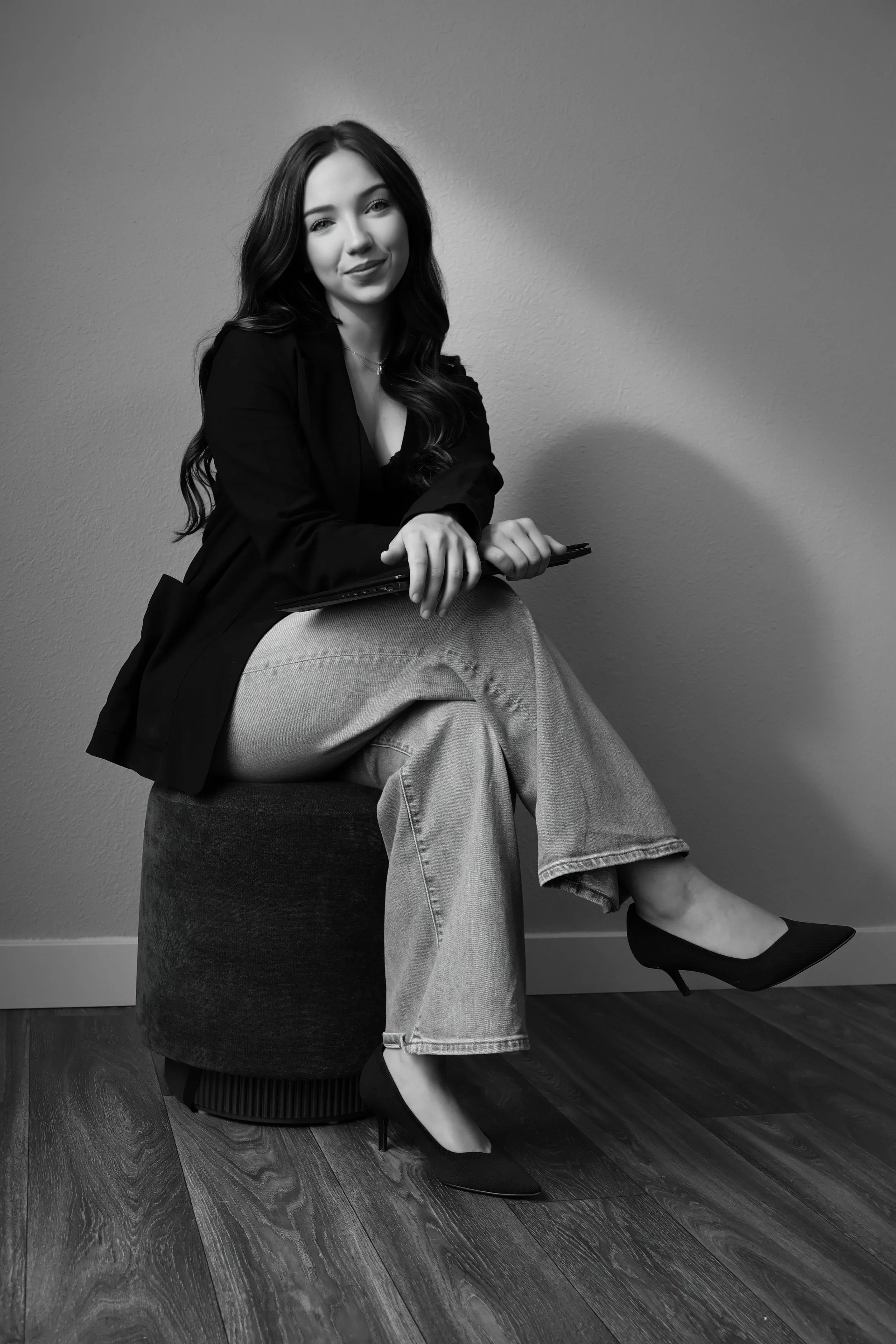 Black and white photo of a woman sitting on a round ottoman, dressed in a blazer, a skirt, and high heels, with one leg crossed over the other and holding a closed laptop in her hands.