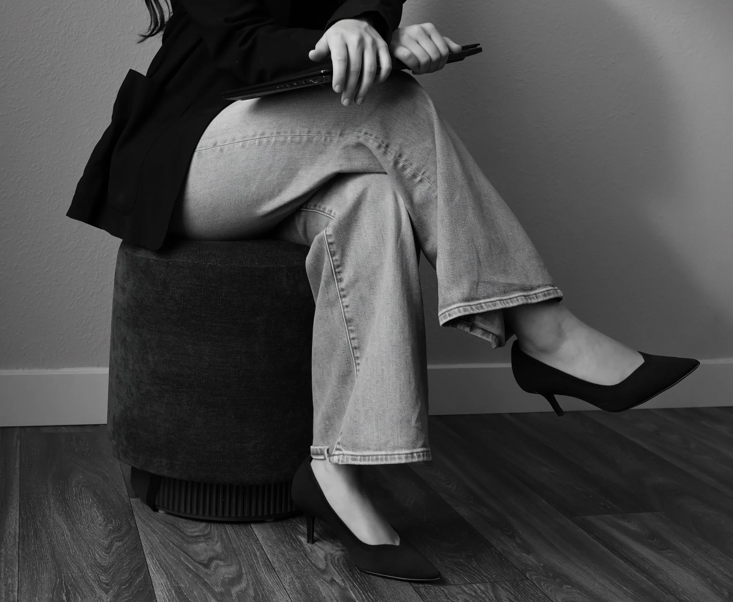A person sitting on a chair against a plain wall, wearing light-colored jeans, a dark top, and black shoes, holding a closed laptop in their hands.