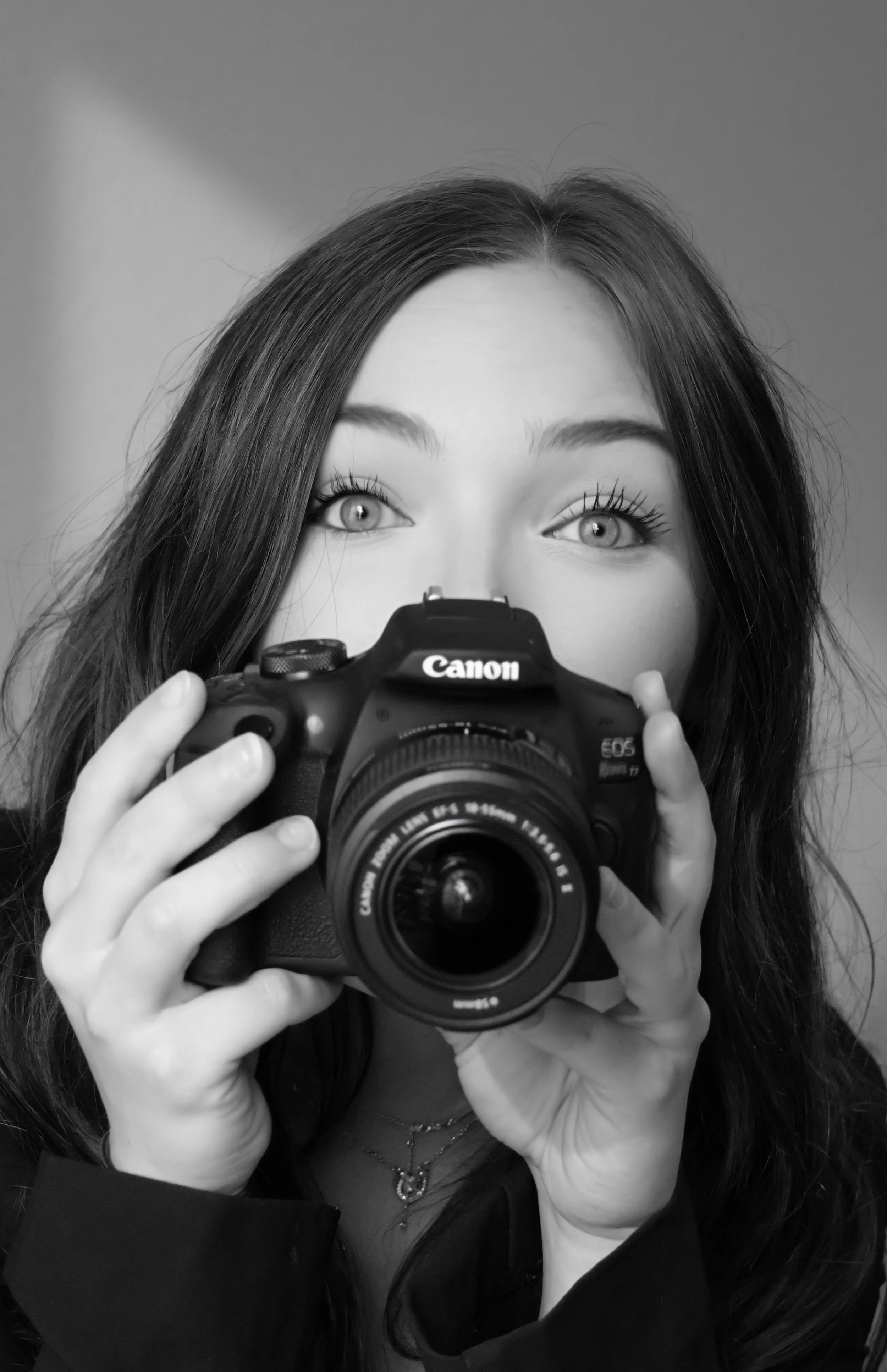 A woman with long dark hair taking a selfie using a Canon camera, with her eyes wide open and focusing on the lens, against a plain background.