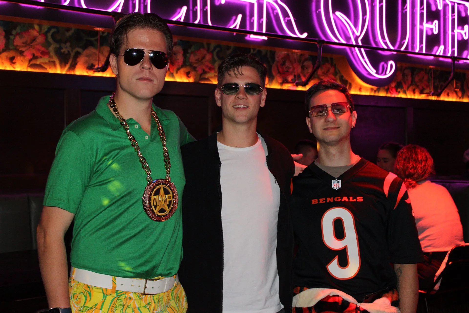 Three men are standing together in a nightclub with neon purple signs and floral decor in the background. The man on the left wears sunglasses, a green polo shirt, yellow patterned shorts, a gold chain, and a large MVP medallion. The man in the middle wears sunglasses, a black jacket over a white T-shirt. The man on the right wears glasses, a black NFL Cincinnati Bengals jersey, and has a red lighting reflection on his face.